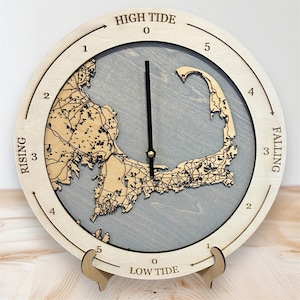 May include: A wooden clock with a map of Cape Cod on the face. The clock has a black hour hand and a gold minute hand. The clock face is divided into 12 sections, with the numbers 1 through 5 on the top half and the numbers 1 through 5 on the bottom half. The clock also has the words "HIGH TIDE" and "LOW TIDE" printed on it.