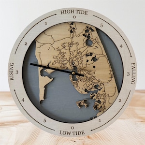 Puede incluir: Un reloj de mareas redondo de madera con un marco de madera clara y un centro gris. La esfera del reloj presenta un diseño de mapa detallado en un tono de madera más claro. Las palabras "HIGH TIDE", "RISING", "FALLING" y "LOW TIDE" están impresas.