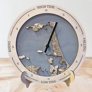May include: A round wooden tide clock with a light blue background and a detailed map design. The clock face displays "HIGH TIDE" and "LOW TIDE" with numbers and arrows indicating rising and falling tides. The clock is supported by a decorative stand.