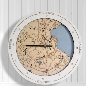 May include: A circular wooden tide clock featuring a laser-cut map of a coastal region with light wood landmasses and a blue water section. The white outer ring displays the text HIGH TIDE, LOW TIDE, RISING, and FALLING with numerical indicators.