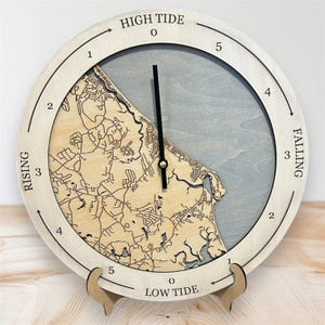 Puede incluir: Un reloj de mareas redondo de madera, con un marco de madera clara y un diseño de mapa. La esfera del reloj muestra las palabras "Marea Alta", "Creciente", "Menguante" y "Marea Baja". El reloj está sostenido por un soporte de madera.