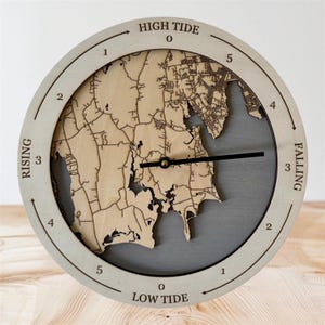 Puede incluir: Un reloj de mareas redondo de madera con diseño de mapa. La esfera del reloj es de madera clara con un borde gris y una manecilla negra. Las palabras "HIGH TIDE", "RISING", "FALLING" y "LOW TIDE" están impresas.