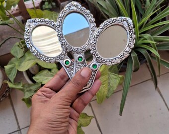 May include: A silver-colored hand mirror with three oval-shaped mirrors. The mirror frames are decorated with floral patterns and green gemstones. The mirror is held in front of a background of green plants.