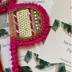 May include: A vibrant pink and gold decorative fan with a tassel fringe. The fan features gold sequins, mirrors, and decorative trim. The text "You're the coolest" is visible on the surrounding cards. The fan is likely used for a special occasion.