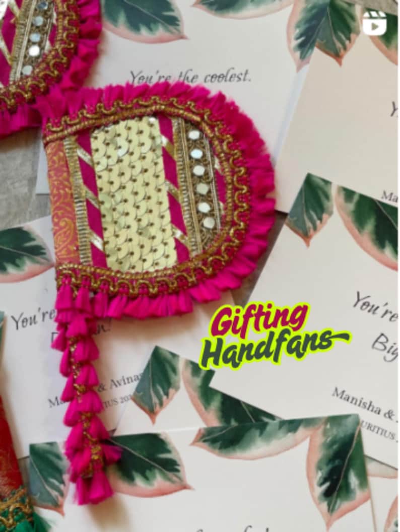May include: Bright pink hand fans with gold trim, sequins, and tassels. The fans are decorated with mirrors and are placed on top of wedding invitation cards. The text "Gifting Handfans" is visible.