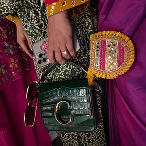 May include: A dark green, crocodile-textured handbag with a gold "C" clasp. The bag is paired with sunglasses and a decorative, fringed accessory. The person is wearing a green patterned outfit with colorful accents and holding a phone.