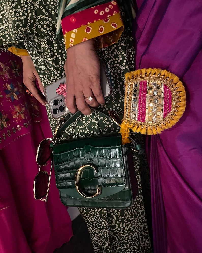 May include: A dark green, crocodile-textured handbag with a gold "C" clasp. The bag is paired with sunglasses and a decorative, fringed accessory. The person is wearing a green patterned outfit with colorful accents and holding a phone.