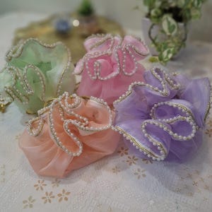 Può includere: Quattro piccole pochette in tessuto arricciato nelle tonalità pastello verde menta, rosa tenue, pesca e lavanda. Ogni pochette presenta un bordo a volant rifinito con un filo metallico dorato e una delicata bordura di piccole perle bianche.