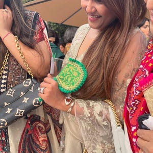 May include: A green and gold clutch with a scalloped edge is held by a person. The clutch has a gold chain strap. The person is wearing a gold-colored embroidered top and a silver bracelet. A black handbag with a gold chain strap is also visible.