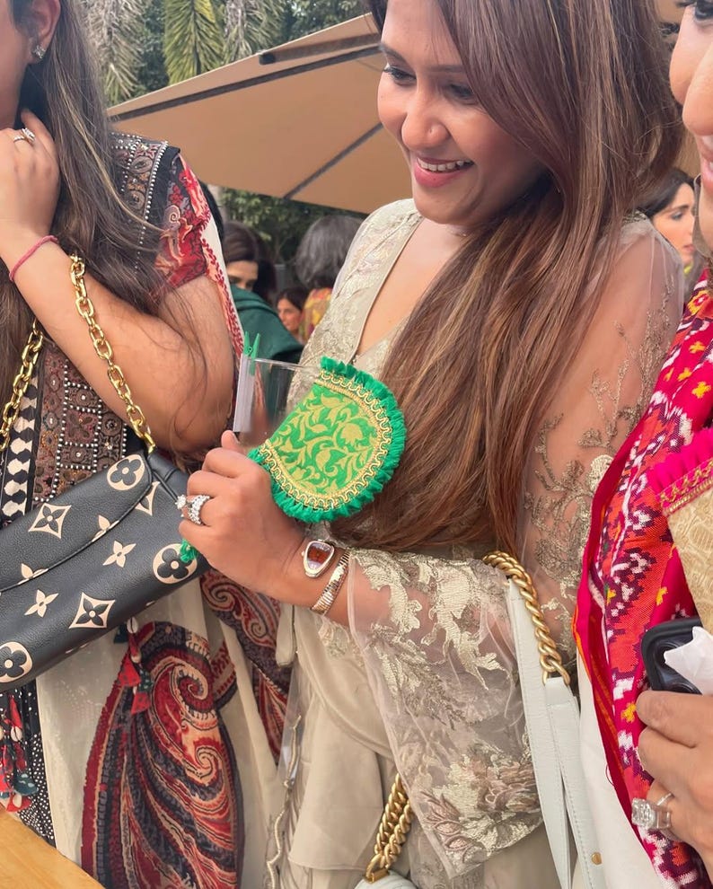 May include: A green and gold clutch with a scalloped edge is held by a person. The clutch has a gold chain strap. The person is wearing a gold-colored embroidered top and a silver bracelet. A black handbag with a gold chain strap is also visible.
