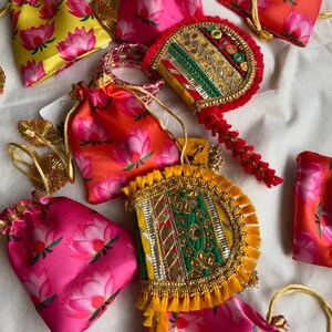 May include: An assortment of colorful, decorative pouches and embellished fans. The pouches are made of satin and feature a pink lotus flower pattern on a yellow, pink, and orange background. The fans are adorned with gold trim and colorful embellishments.