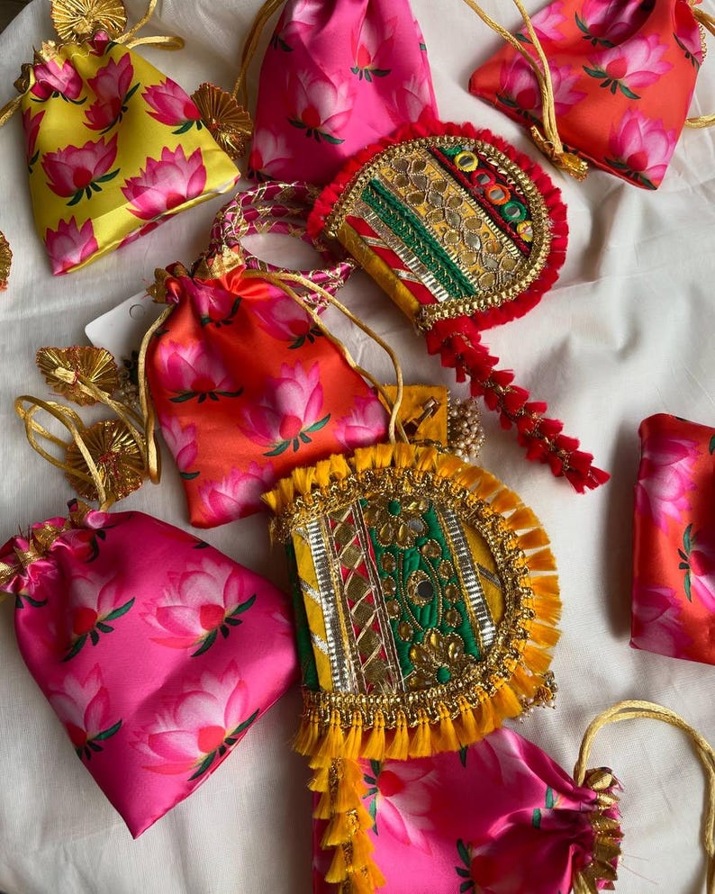 May include: An assortment of colorful, decorative pouches and embellished fans. The pouches are made of satin and feature a pink lotus flower pattern on a yellow, pink, and orange background. The fans are adorned with gold trim and colorful embellishments.