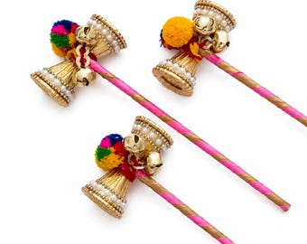 May include: Three decorative sticks featuring gold-toned bell-shaped ornaments adorned with white faux pearls, small metallic jingle bells, and vibrant multi-colored pom-poms. Each stick is wrapped in a pink and gold striped ribbon pattern.