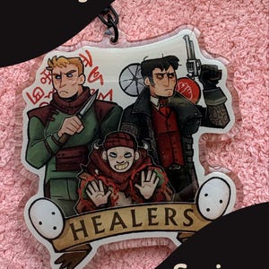 May include: A clear acrylic keychain featuring three illustrated characters from Pathologic. One holds a knife, another a revolver, and a third figure is centered below. The design includes red symbols, a banner with the text HEALERS, and two white masks.