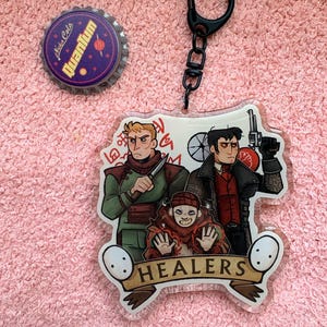 May include: An acrylic keychain featuring three illustrated characters and the text HEALERS, measuring 2.5 inches wide by 2.75 inches high, accompanied by a circular Nuka Cola Quantum bottle cap with a purple background and yellow text.
