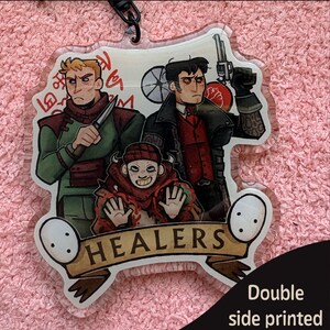 May include: A clear acrylic keychain featuring a double-sided epoxy finish with an illustration of three characters. Two figures stand behind a smaller character with a banner at the bottom that reads HEALERS. The text Double sided epoxy and Double side printed Same is visible.