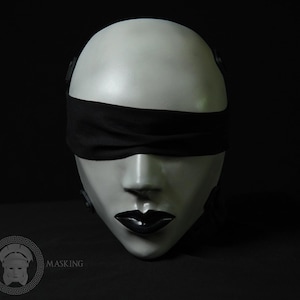 May include: A white mask with a black blindfold covering the eyes and black lipstick. The mask has a smooth, pale finish and is set against a dark background. The mask is a theatrical or costume piece.