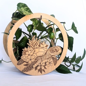 Puede incluir: Un soporte de madera para plantas con un diseño cortado con láser de una tortuga marina adornada con flores. La tortuga está centrada dentro de un marco circular, con un segundo anillo de madera más grande detrás. Plantas verdes llenan el espacio.