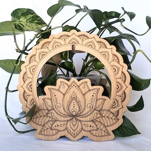 May include: A wooden plant holder with a laser-cut lotus flower design. The holder has a circular frame with a scalloped edge and intricate patterns. Green leafy plants are visible behind the holder, creating a natural aesthetic.