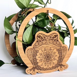 Puede incluir: Un soporte de madera para plantas con un diseño de rana cortado con láser. La rana está decorada con un patrón de mandala. El soporte tiene un marco circular y está lleno de plantas verdes.
