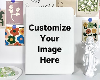 Op de afbeelding: Een wit notitieboek met de tekst "Customize Your Image Here" in het zwart. Het notitieboek ligt op een wit oppervlak, met decoratieve kaarten en een klein engelenbeeldje op de achtergrond.