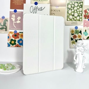 Op de afbeelding: Een witte tablethoes met een textuuroppervlak. Het woord "Office" is zichtbaar op een kaart. Decoratieve items zijn een klein engelenbeeld en kleurrijke bloemenkunstprints.