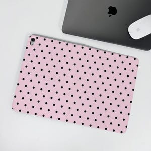 Op de afbeelding: Een roze tablethoes met een patroon van gelijkmatig verdeelde zwarte stippen. De hoes is open en toont het tabletscherm. Een laptop en muis zijn op de achtergrond te zien.