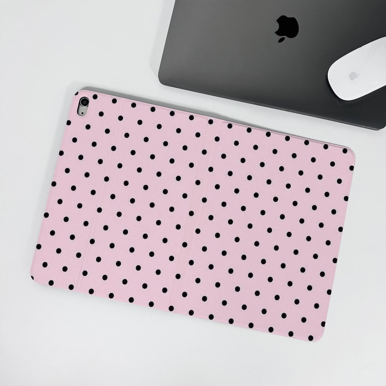 Op de afbeelding: Een roze tablethoes met een patroon van gelijkmatig verdeelde zwarte stippen. De hoes is open en toont het tabletscherm. Een laptop en muis zijn op de achtergrond te zien.