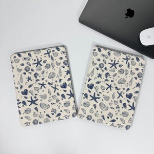 Può includere: Due custodie per tablet color crema con un motivo ripetuto di stelle marine e conchiglie blu navy. Le custodie sono rettangolari con angoli arrotondati. Un laptop e un mouse sono visibili sullo sfondo.