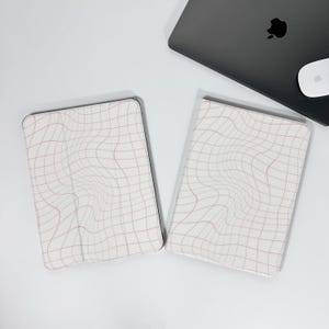 Könnte beinhalten: Zwei weiße Tablet-Hüllen mit einem rosa Gittermuster und ein schwarzes Laptop mit einer weißen Maus auf einer weißen Oberfläche. Das Gittermuster scheint verzerrt zu sein und erzeugt einen wellenförmigen Effekt.