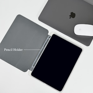 Op de afbeelding: Een grijze tablethoes met een transparante rand, open om een zwart scherm en een potloodhouder te onthullen. Een grijze laptop en een witte muis staan op de achtergrond. De tekst "Pencil Holder" is zichtbaar op de hoes.