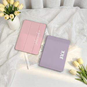 May include: Two tablet cases, one pink and one lavender, are displayed on a white surface. The pink case has the name "John Smith" in white script, and the lavender case has the name "JACK" in white block letters. The cases are surrounded by white fabric and flowers.