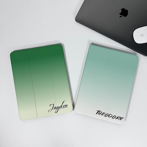 Op de afbeelding: Twee tablethoezen met een gradiëntontwerp. Eén hoes is groen, vervaagt naar wit, met de naam "Jayden" in script. De andere heeft een turquoise gradiënt naar wit, met de naam "THEODORE" in script. Een laptop en muis staan op de achtergrond.