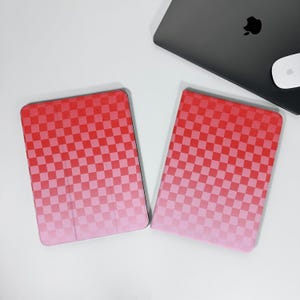 Può includere: Due custodie per tablet a scacchi rossi e rosa sono esposte su una superficie bianca. Le custodie hanno un effetto sfumato, che passa dal rosso al rosa. Un laptop grigio e un mouse bianco sono sullo sfondo.