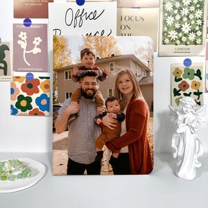May include: A rectangular photo print featuring a family of four, with a man carrying a child on his shoulders and a woman holding a baby. The print is surrounded by decorative cards and a white angel statue.
