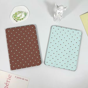 Può includere: Due custodie per tablet con motivi a cuore. Una custodia è marrone con cuori bianchi, l'altra è azzurra con cuori marroni. Sono visibili anche una piccola statuina di angelo bianco e un piattino con gioielli verdi.