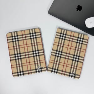 Könnte beinhalten: Zwei beige Tablet-Hüllen mit einem klassischen Karomuster in Rot, Schwarz und Weiß. Die Hüllen haben abgerundete Ecken und eine personalisierte Schrift. Ein Laptop und eine Maus befinden sich im Hintergrund.