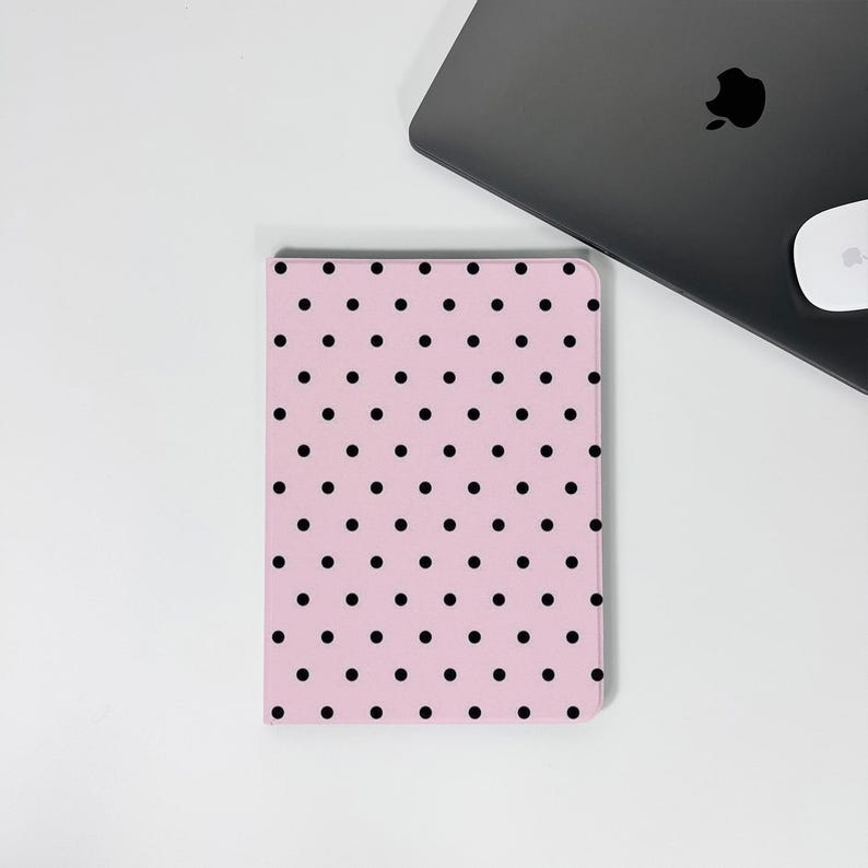 Op de afbeelding: Een roze notitieboek met een zwart stippenpatroon. Het rechthoekige notitieboek ligt op een wit oppervlak, naast een gesloten grijze laptop en een witte muis.