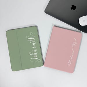 Può includere: Due custodie per tablet, una verde salvia e una rosa, sono esposte su una superficie bianca. La custodia verde salvia ha il nome "John Smith" in corsivo bianco e la custodia rosa ha "Nathaniel Neil" in corsivo bianco. Un laptop e un mouse sono sullo sfondo.