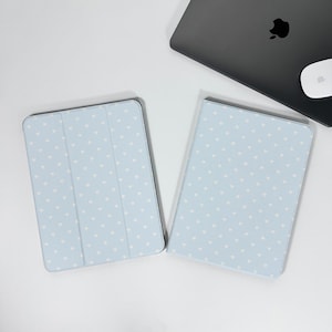 May include: Two light blue tablet cases with a white heart pattern are displayed on a white surface. A closed black laptop and a white mouse are in the background. The cases are designed to protect tablets.