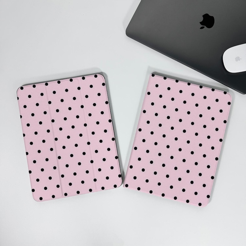 Op de afbeelding: Twee roze tablethoezen met een zwart stippenpatroon. E&eacute;n hoes is open en onthult de binnenkant, terwijl de andere gesloten is. Een laptop en muis staan op de achtergrond.