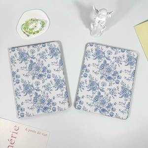 May include: Two tablet cases with a white and blue floral pattern. The cases are rectangular with rounded corners. The floral design features roses and other flowers. The cases are on a white surface with other decorative items.