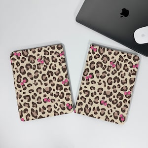 Puede incluir: Dos fundas para tabletas con un estampado de leopardo beige, con manchas marrones y detalles de lazos rosas. Las fundas son rectangulares con esquinas redondeadas. Un portátil y un ratón están en el fondo.