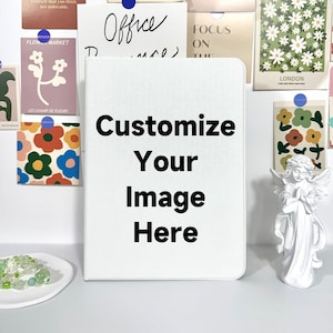Könnte beinhalten: Ein weißes Notizbuch mit dem Text "Customize Your Image Here" in fetten schwarzen Buchstaben. Das Notizbuch ist von dekorativen Gegenständen umgeben, darunter florale Kunstdrucke, eine kleine Engelsstatue und ein Perlenarmband.