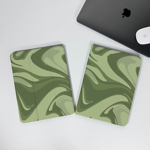 May include: Two olive green tablet cases with a swirling pattern on a white surface. The cases are rectangular with rounded corners. A black laptop and white mouse are in the background.