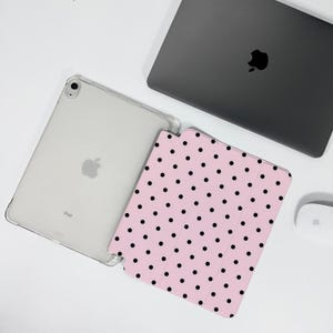Op de afbeelding: Een doorzichtige iPad-hoes, een roze iPad-hoes met zwarte stippen, een grijze laptop en een witte muis zijn op een wit oppervlak geplaatst. De iPad-hoes is transparant en de hoes heeft een patroon van zwarte stippen. De laptop is gesloten en de muis is draadloos.