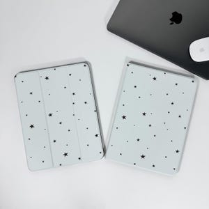 May include: Two light blue tablet cases with a black star pattern. A closed gray laptop and a white mouse are in the background. The cases are rectangular and have a smooth surface.