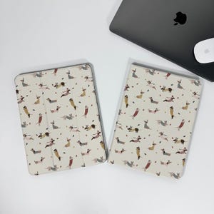 Puede incluir: Dos fundas de tableta color crema con un patrón repetido de perros salchicha en varias poses y atuendos. Un ordenador portátil y un ratón están en el fondo. Las fundas están abiertas, revelando el interior.