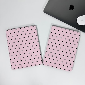 Puede incluir: Dos fundas rosas para tablet con lunares negros. Una funda está abierta, revelando el interior. Un portátil cerrado y un ratón blanco están en el fondo. Las fundas están sobre una superficie blanca.