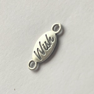 May include: A silver-toned metal connector charm in an elongated oval shape featuring the word Wish embossed in a cursive script. The charm includes a small circular loop at each end for attachment, providing a functional component for jewellery.